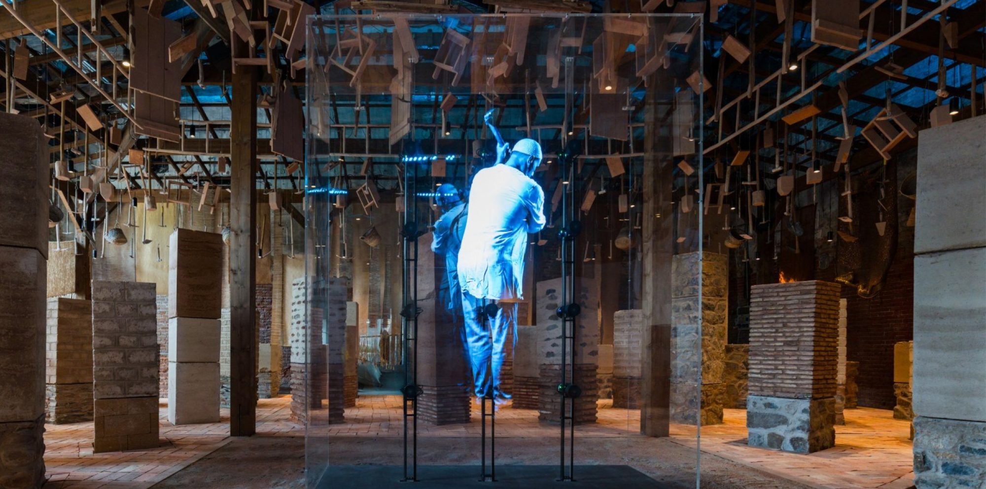 A holographic figure of a craftsman inside a glass case, in a hall of rammed-earth, brick and stone pillars under a ceiling of suspended wooden tools and ladders